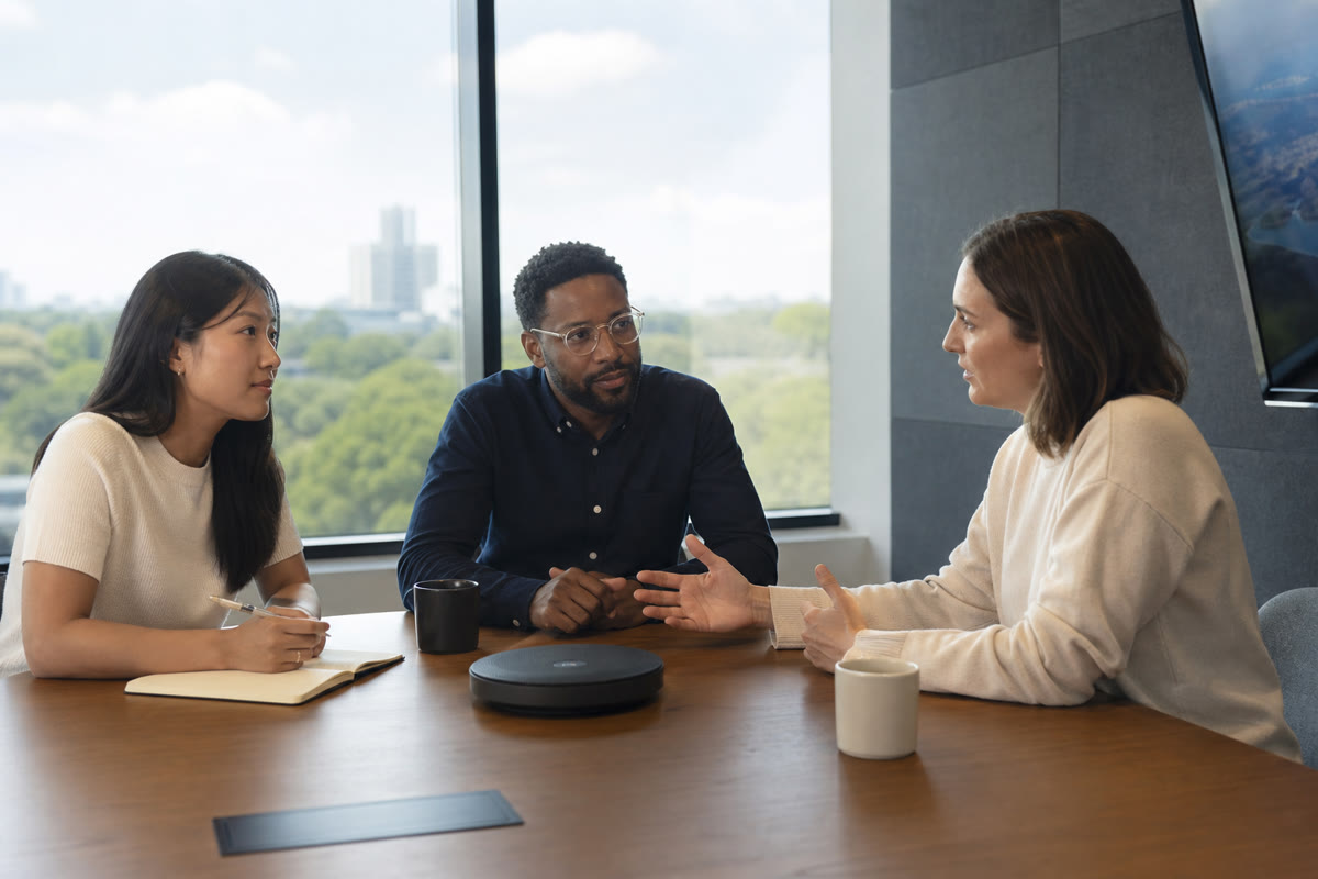 Three colleagues around a conference table talking to a remote participant through a table speakerphone.